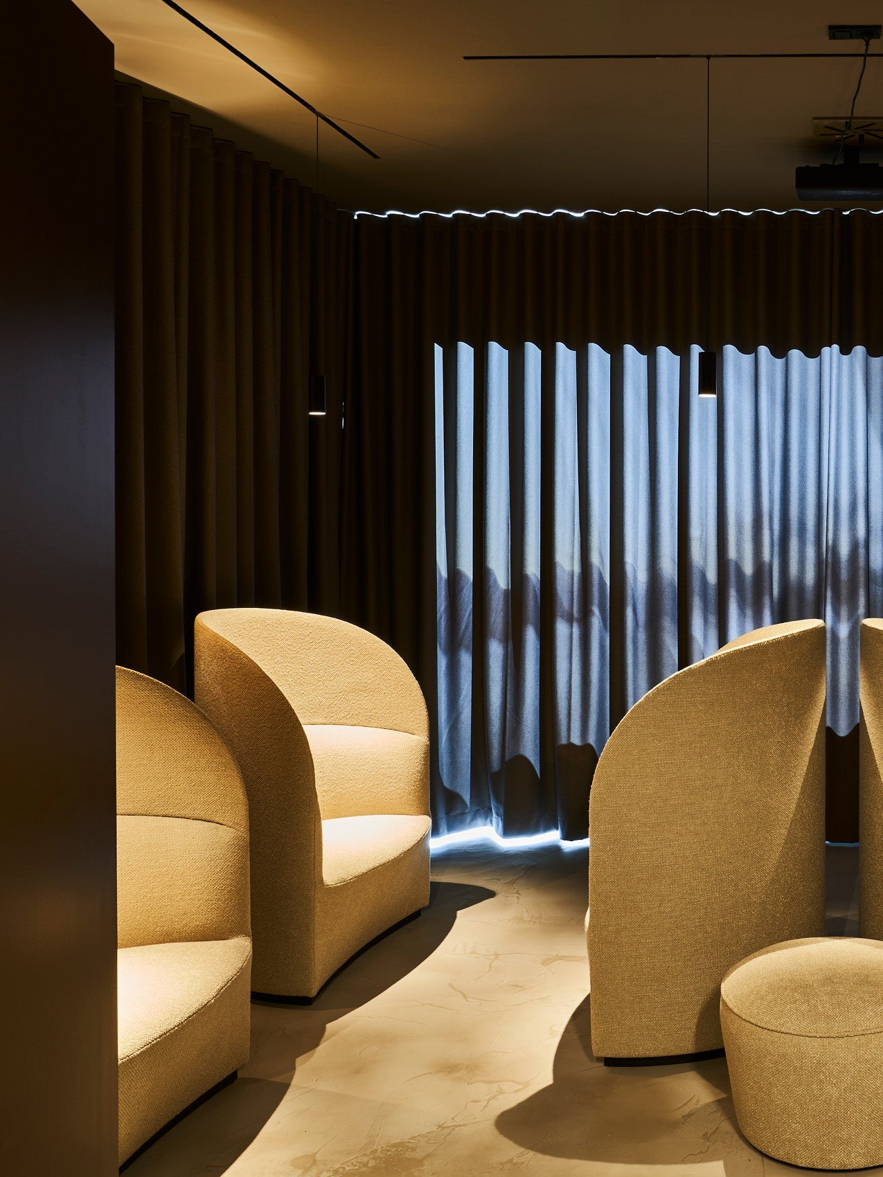 Curved beige upholstered lounge chairs and ottoman in a dim room with dark pleated curtains and soft overhead lighting