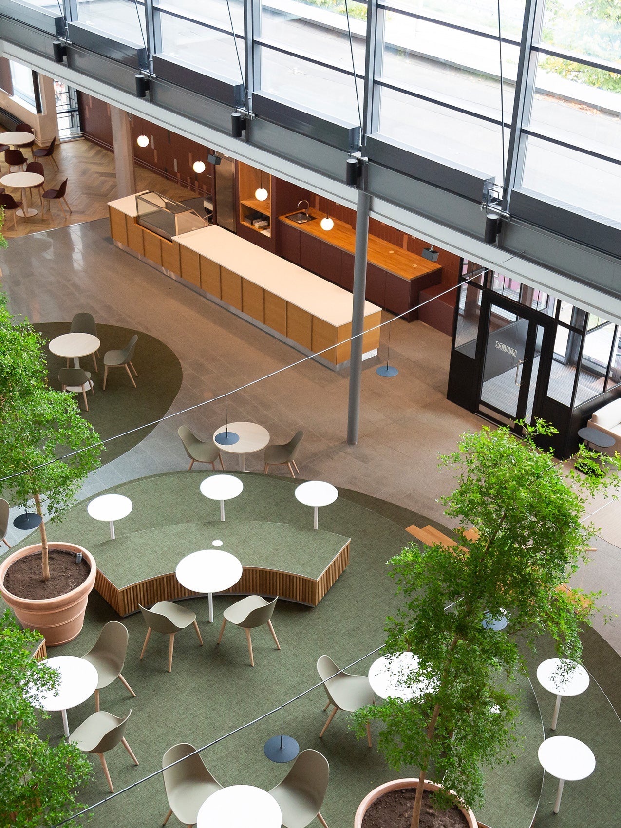 High atrium café with green carpeted circular seating, white round tables, muted olive chairs and large potted trees.