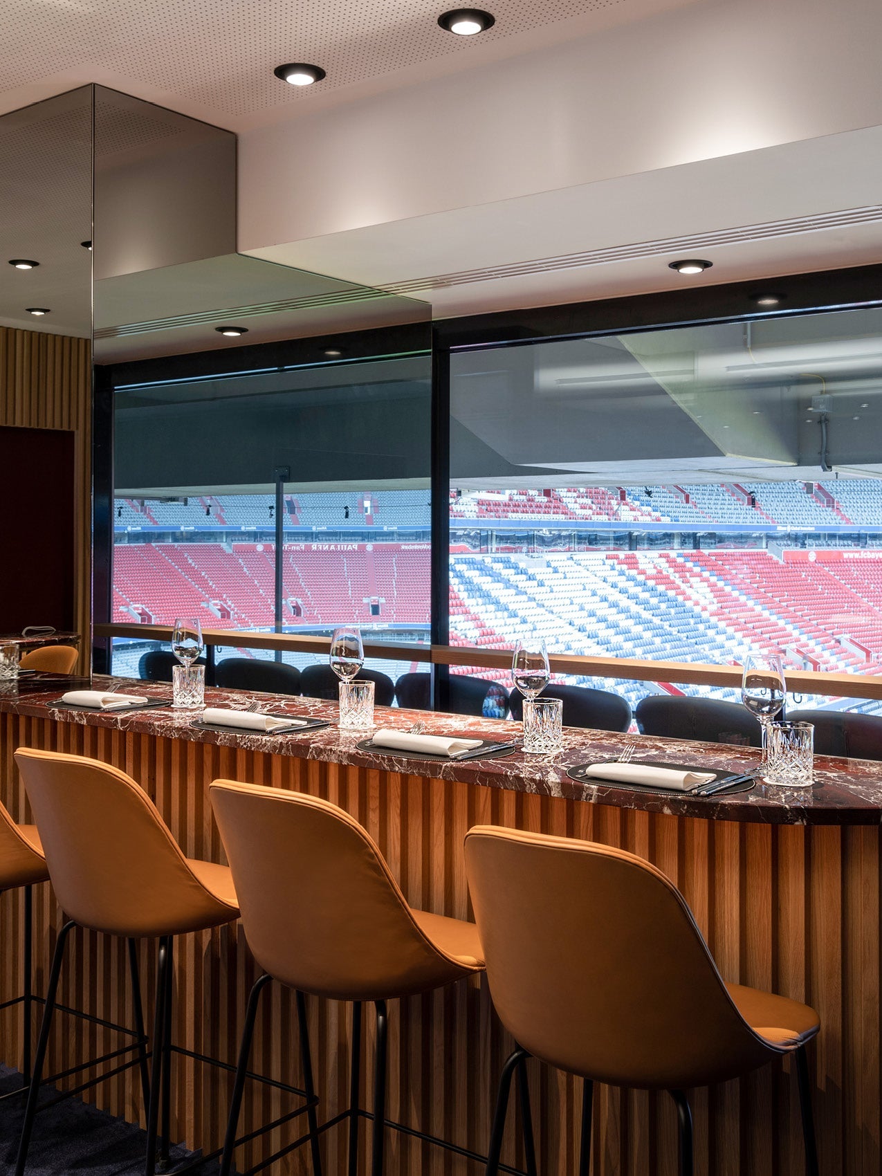Marble-top bar with ribbed wood front, four tan leather high stools, glassware and stadium view through large window