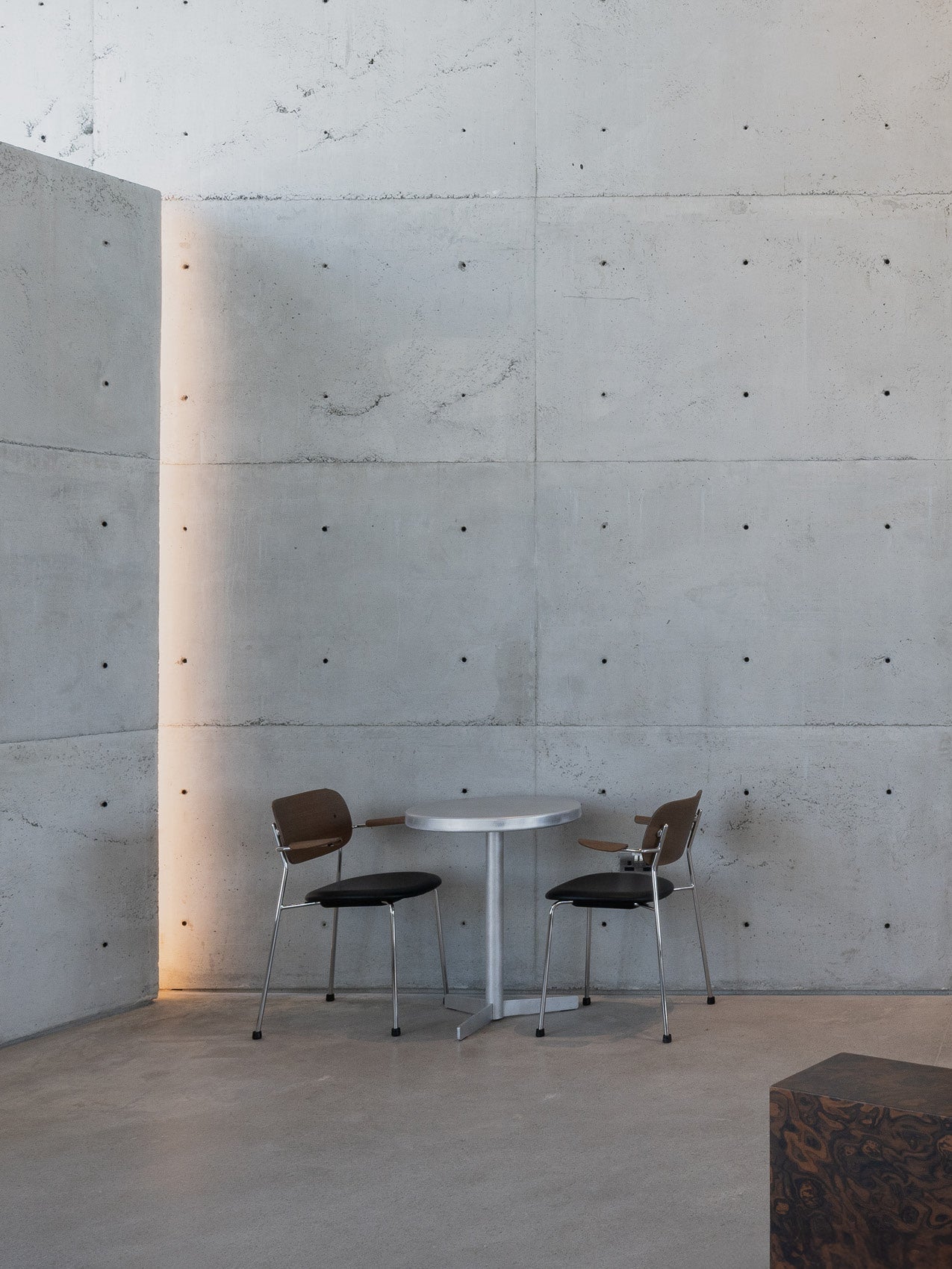 Minimal round metal table with two brown wooden and black leather chairs against a raw concrete wall with vertical light