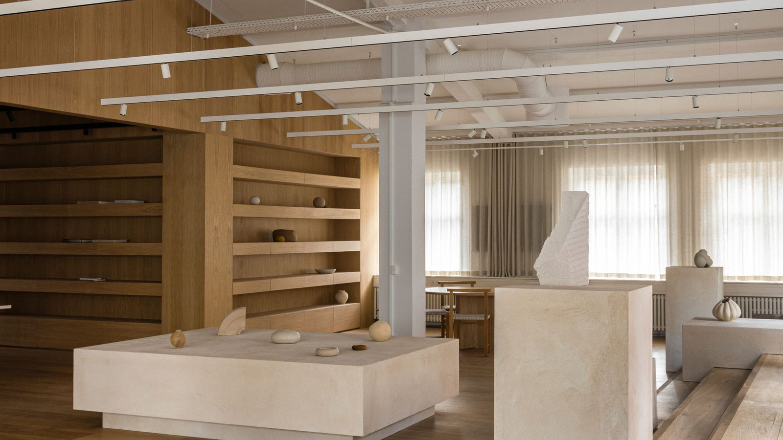 Minimalist showroom interior with pale beige plaster plinths, wooden shelving, neutral ceramic decor and soft filtered