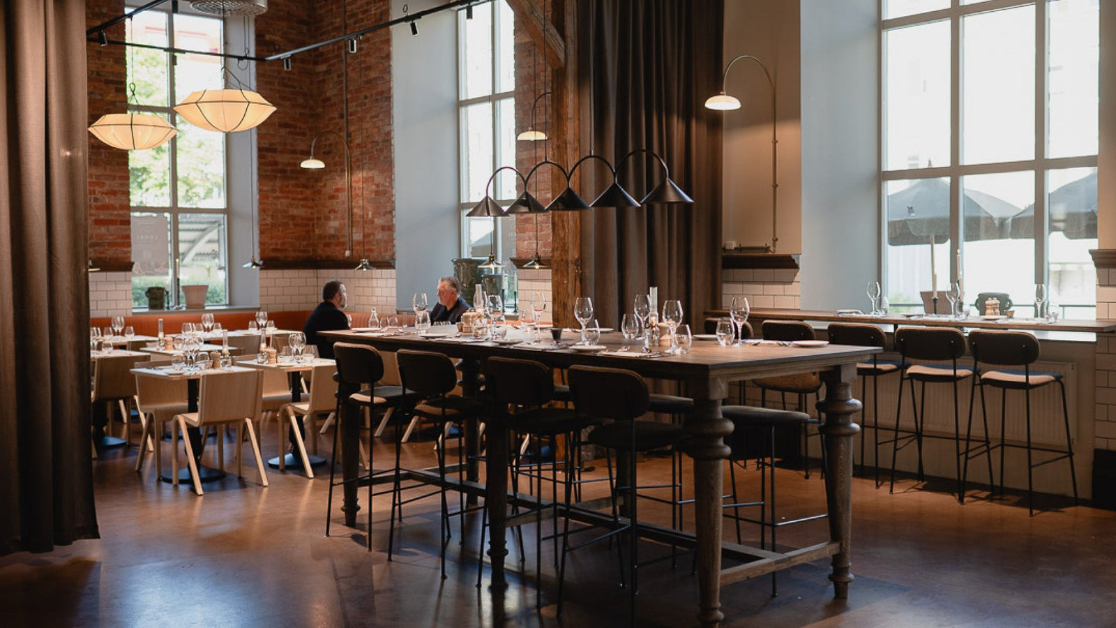 Industrial dining room with wooden high table, black metal bar stools, pendant and arc lamps, exposed brick and large windows
