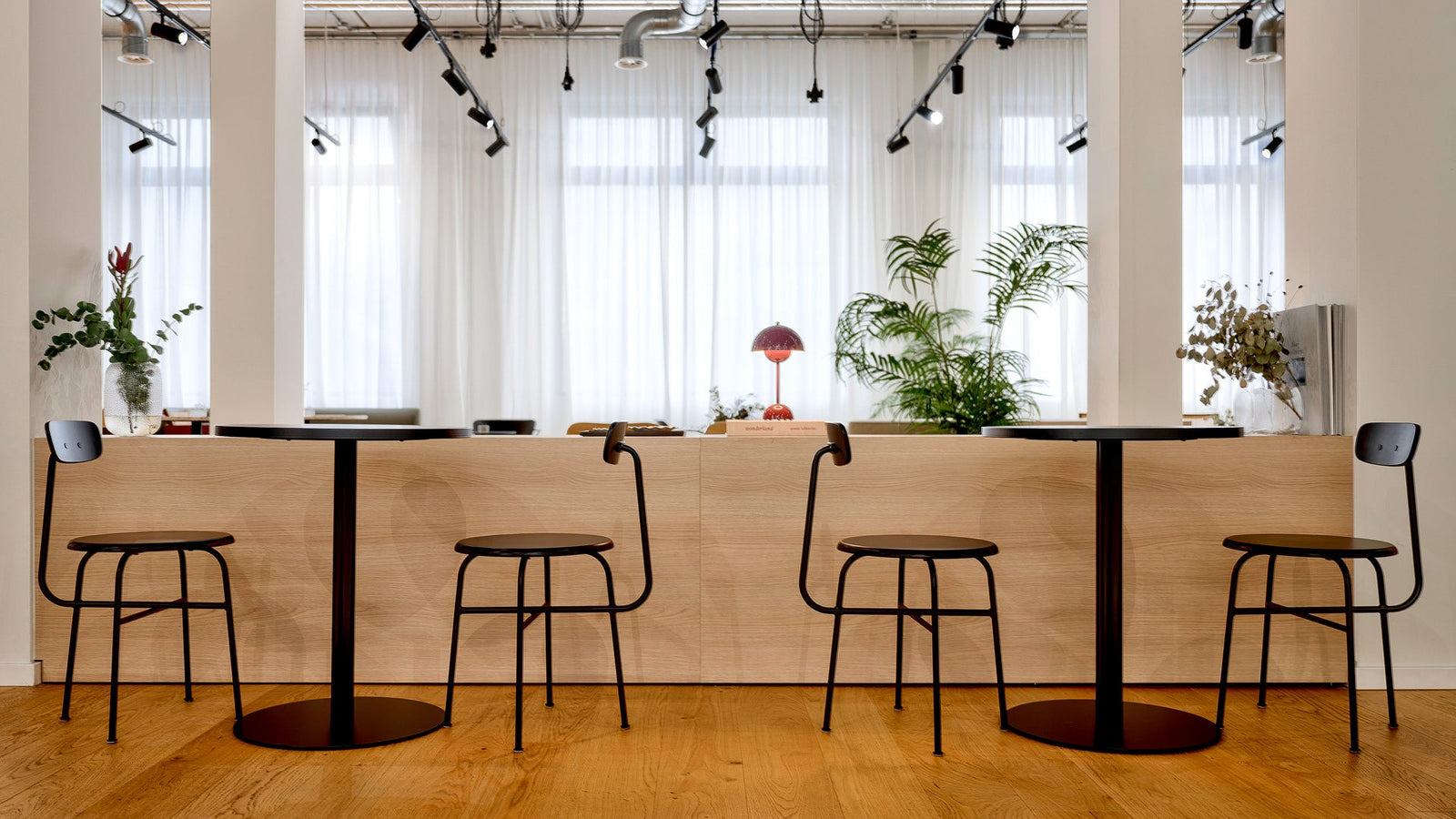 Minimal cafe counter with light oak panel, black metal stools and round tables, glass vases and a red dome table lamp