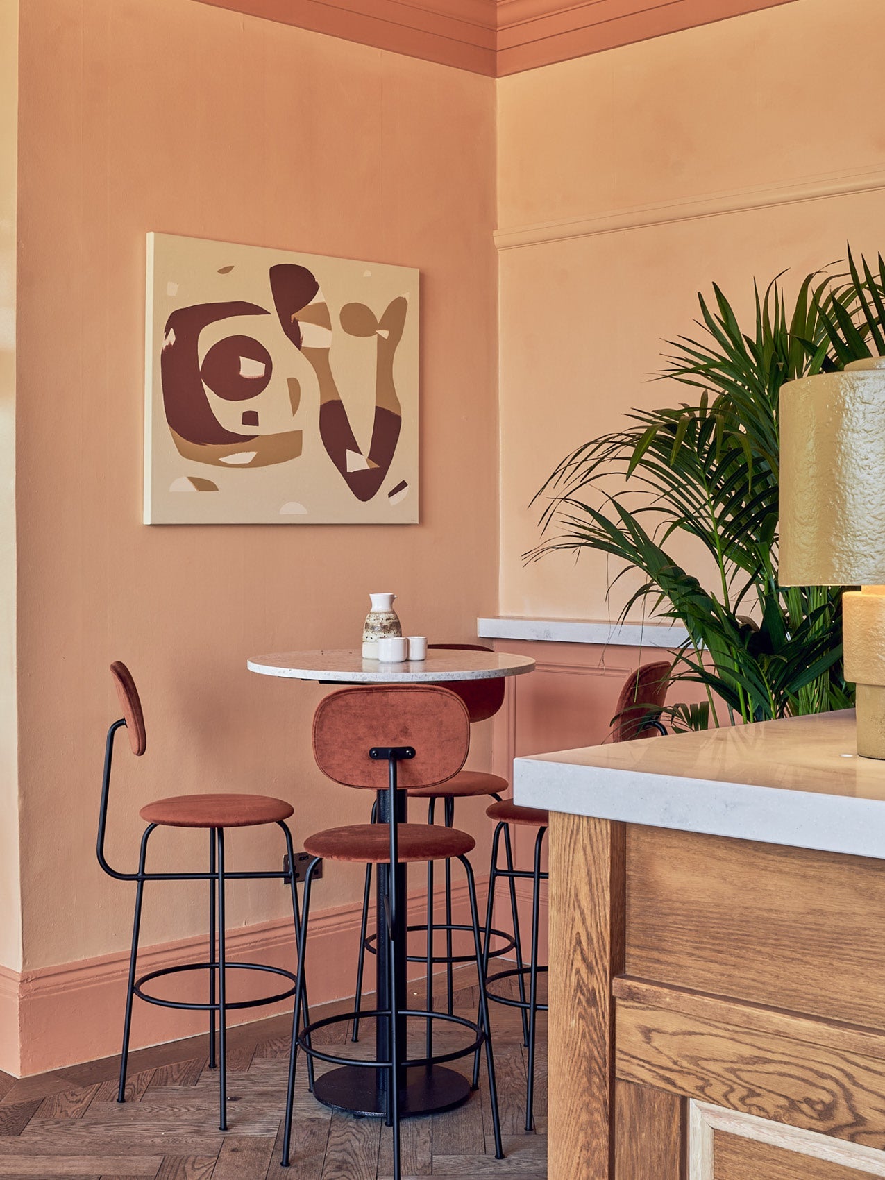 Small café corner with round white marble table, rust velvet bar stools on black metal frames, peach walls and abstract wall