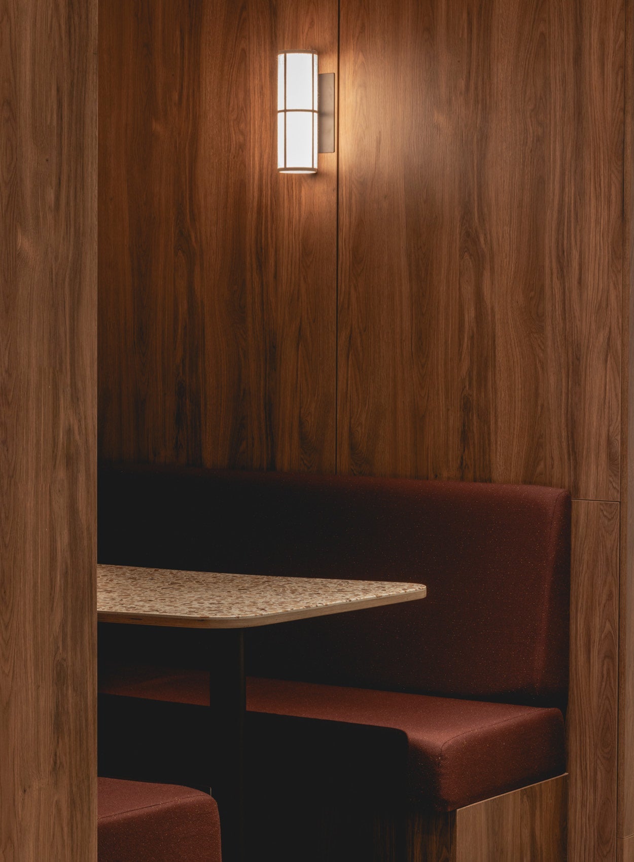 Intimate booth with walnut wood panelling, terrazzo table and burgundy upholstered bench, lit by a cylindrical wall sconce