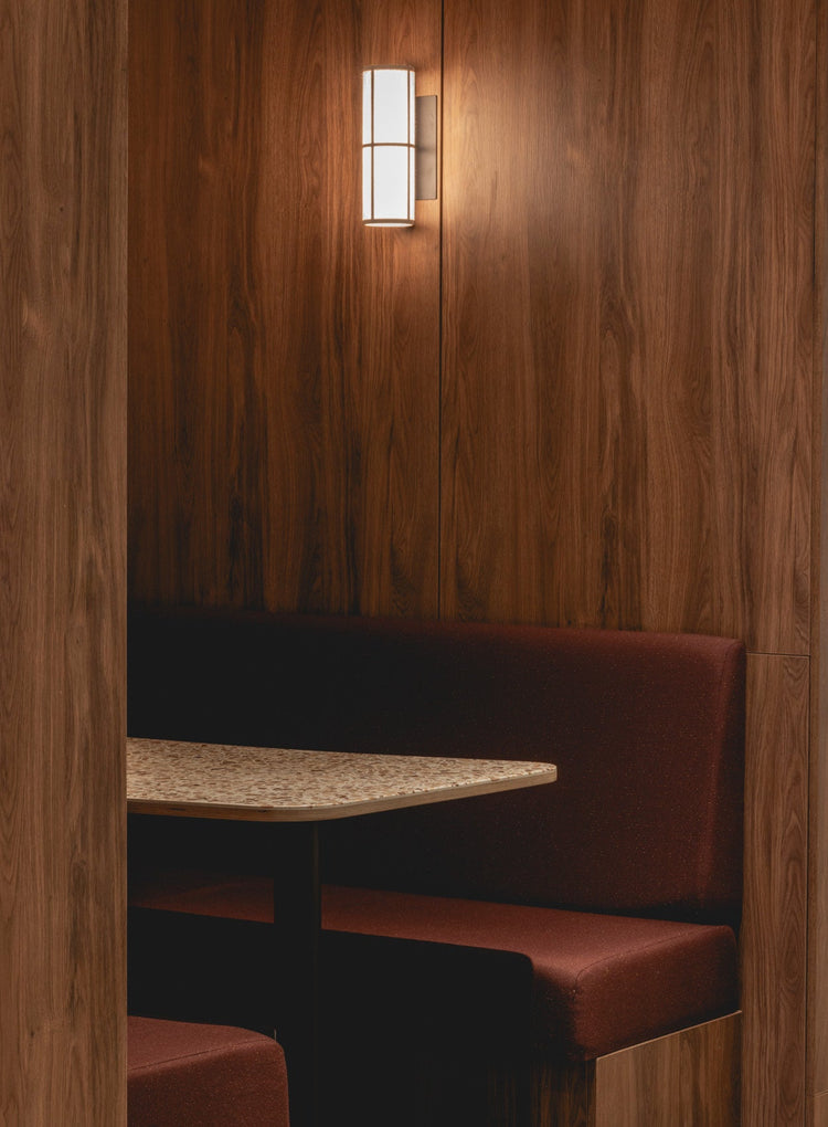 Intimate booth with walnut wood panelling, terrazzo table and burgundy upholstered bench, lit by a cylindrical wall sconce