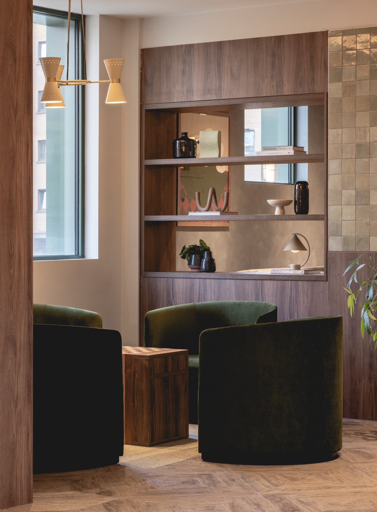 Cozy seating nook with two dark green velvet lounge chairs, wooden side table, built-in walnut shelving and pendant lamp