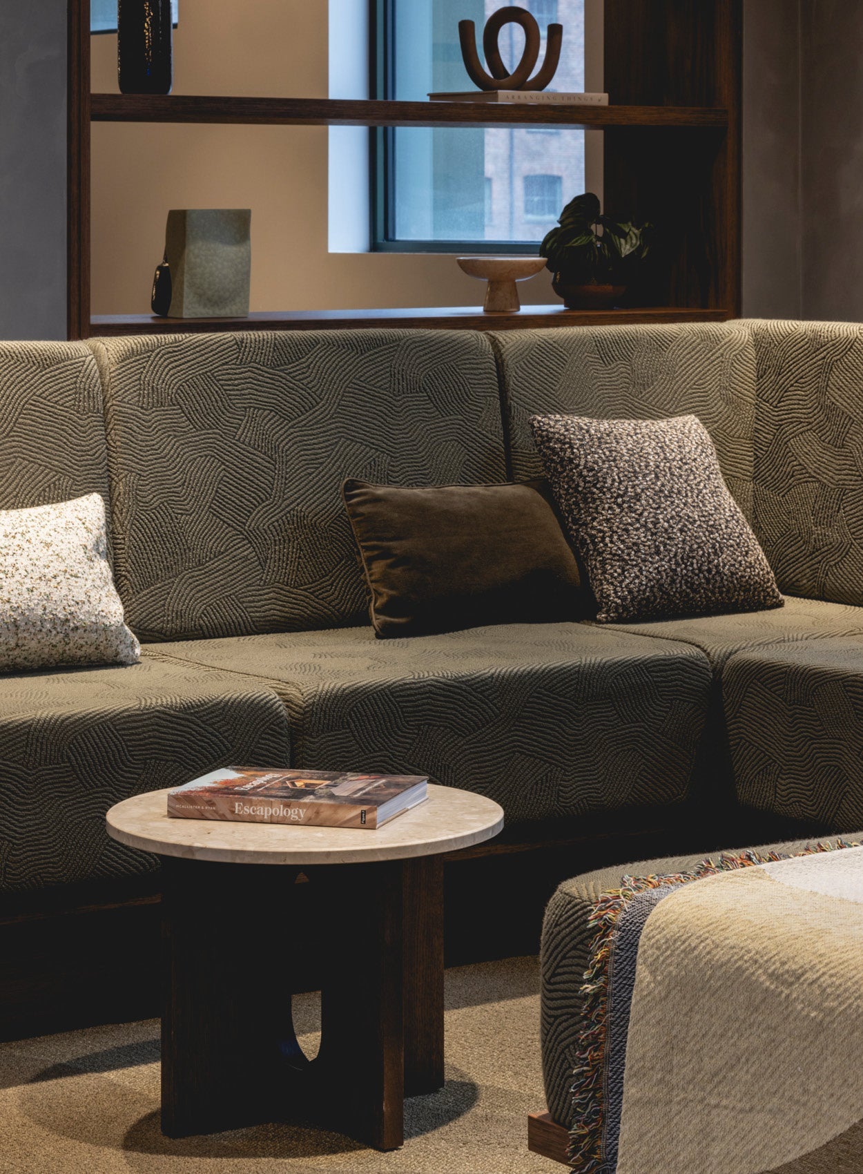 Textured olive-green modular sofa with patterned and velvet cushions, round marble-top side table with book in cozy living