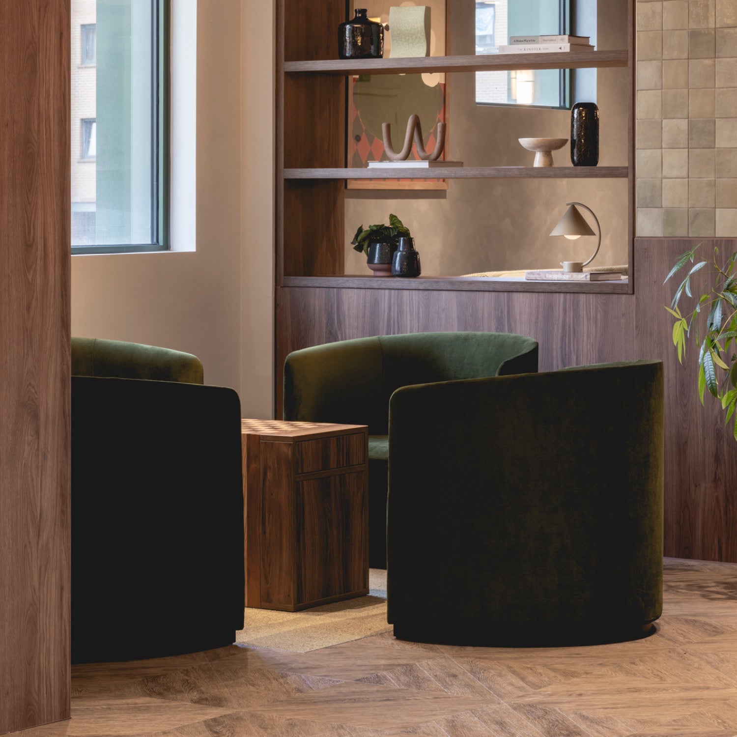 Pair of curved dark green velvet lounge chairs around a wooden cube side table, with walnut shelving and decorative objects