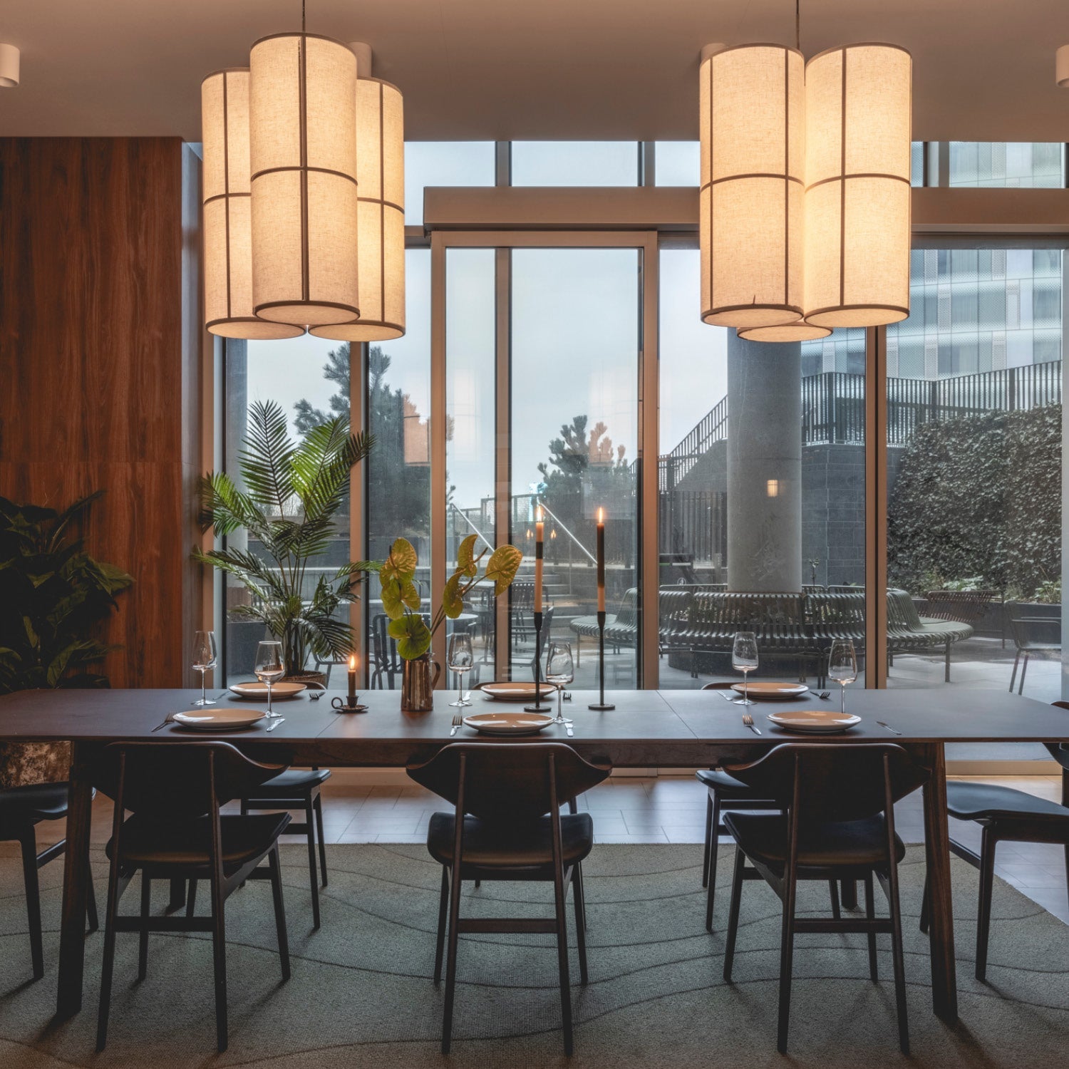 Dining table set with dark wood chairs, tall beige fabric pendant lamps, black candle holders and green potted plants by