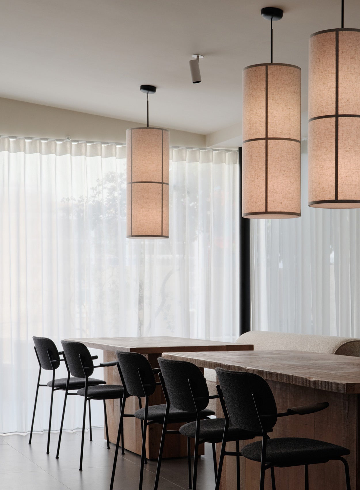 Dining area with long wooden table, black upholstered chairs and three cylindrical beige fabric pendant lamps with dark trim