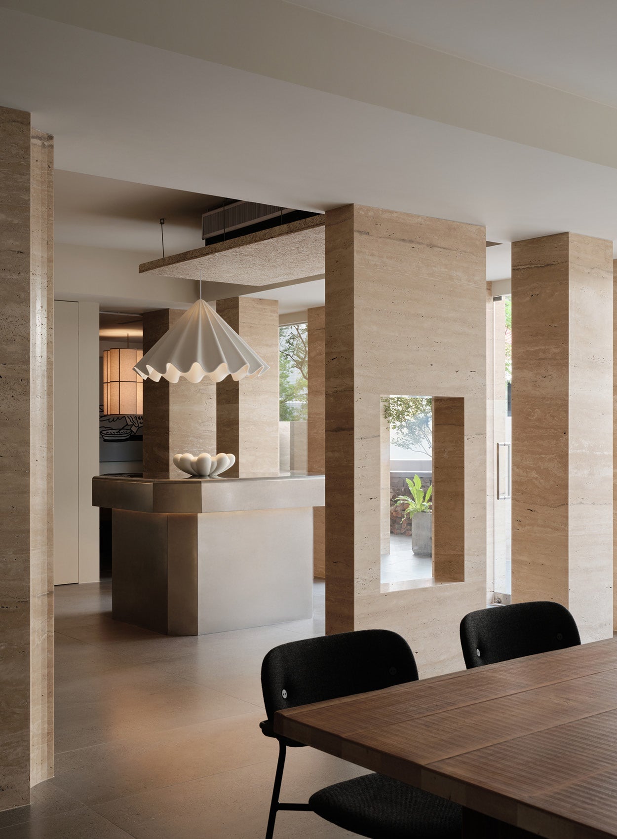 Minimalist dining interior with beige travertine pillars, wooden table, black chairs and white scalloped pendant lamp over