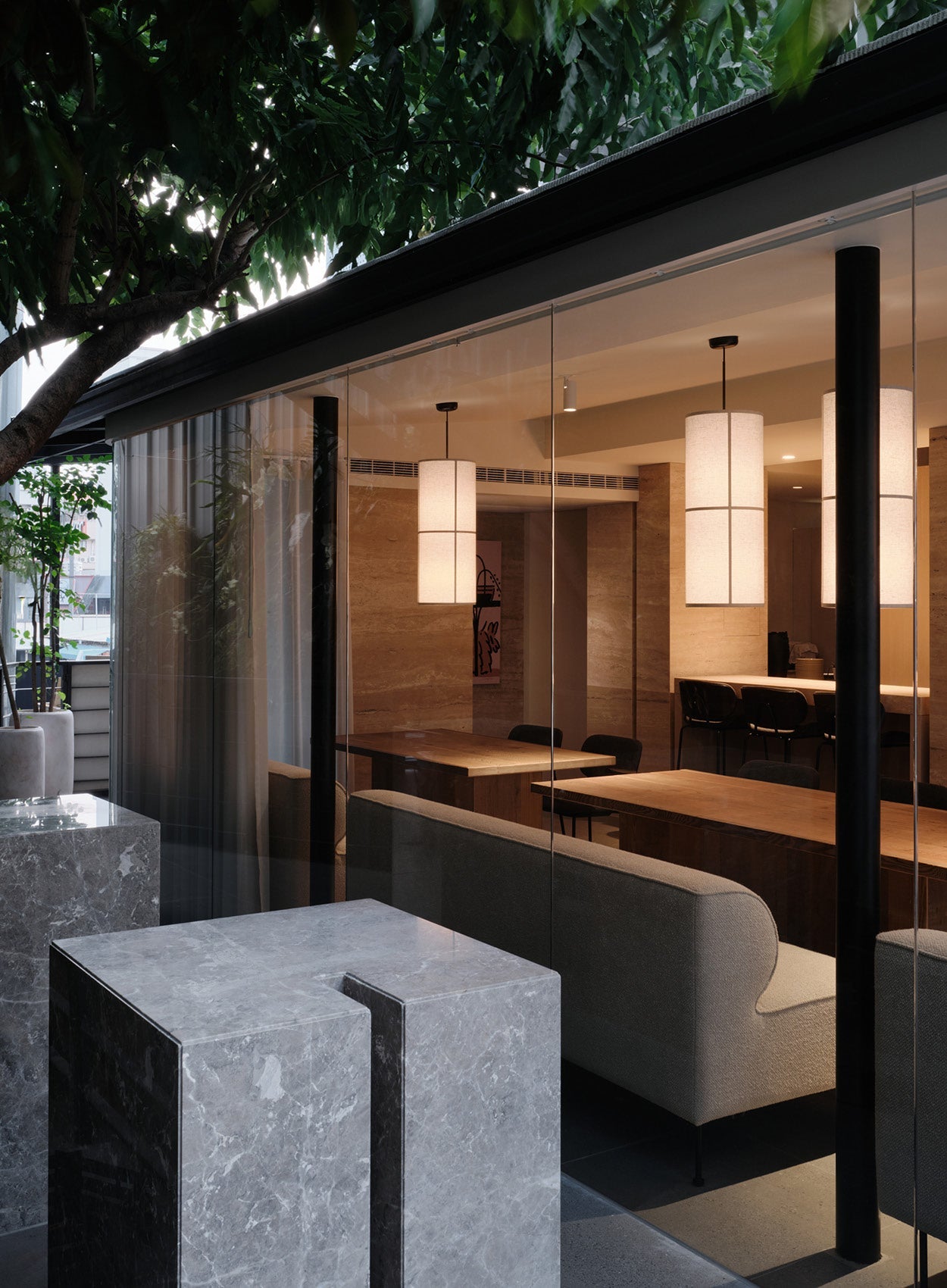 Modern dining interior seen through glass: beige fabric sofas, wooden tables, cylindrical linen pendant lights, grey marble