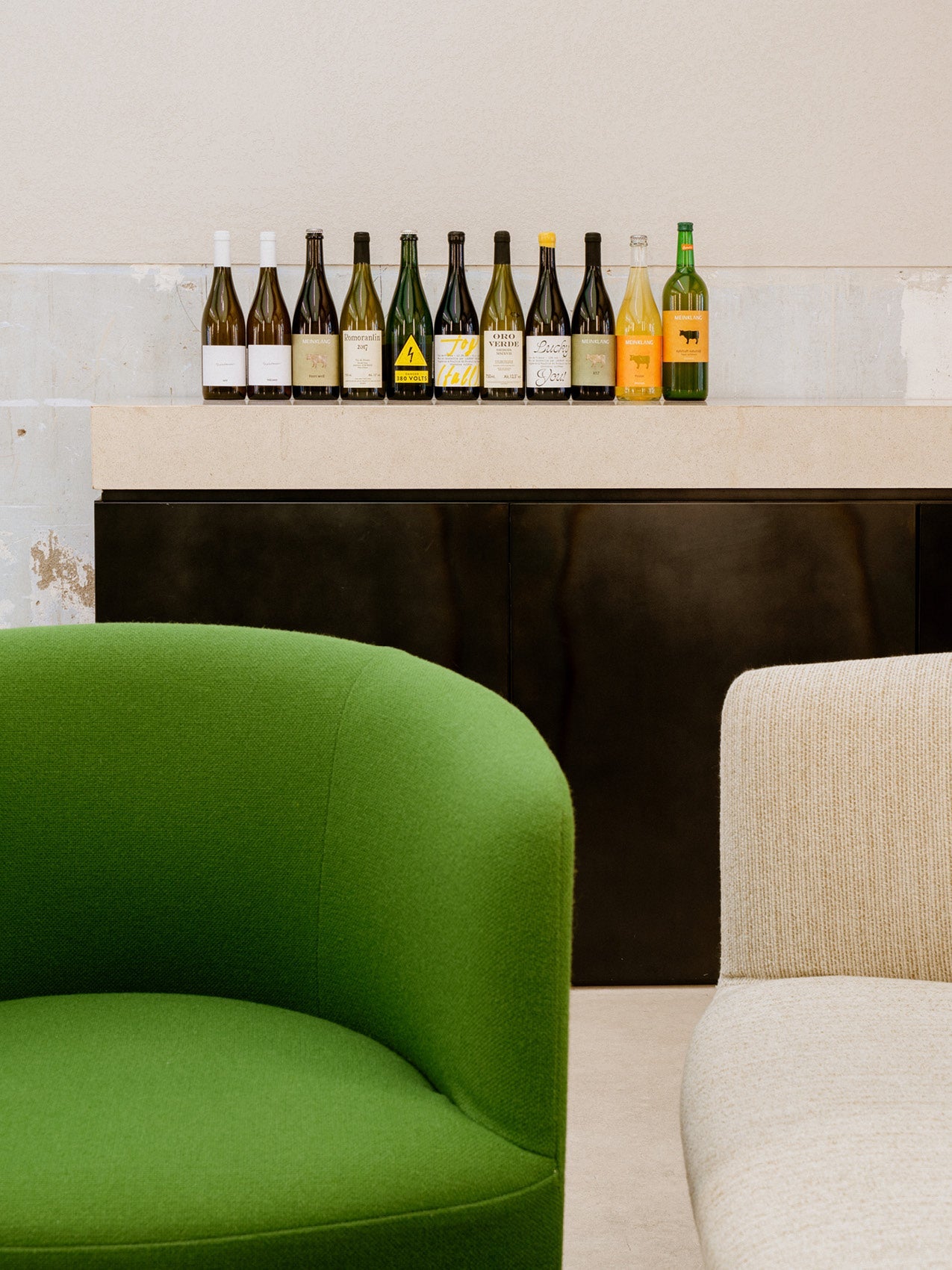Row of assorted green and amber wine bottles on a light stone shelf above black cabinet, foreground curved green and beige