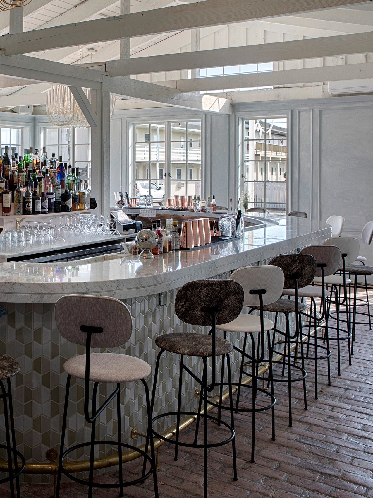 Marble-topped curved bar with hex tile base, row of fabric-upholstered metal barstools, glassware and bottles, bright