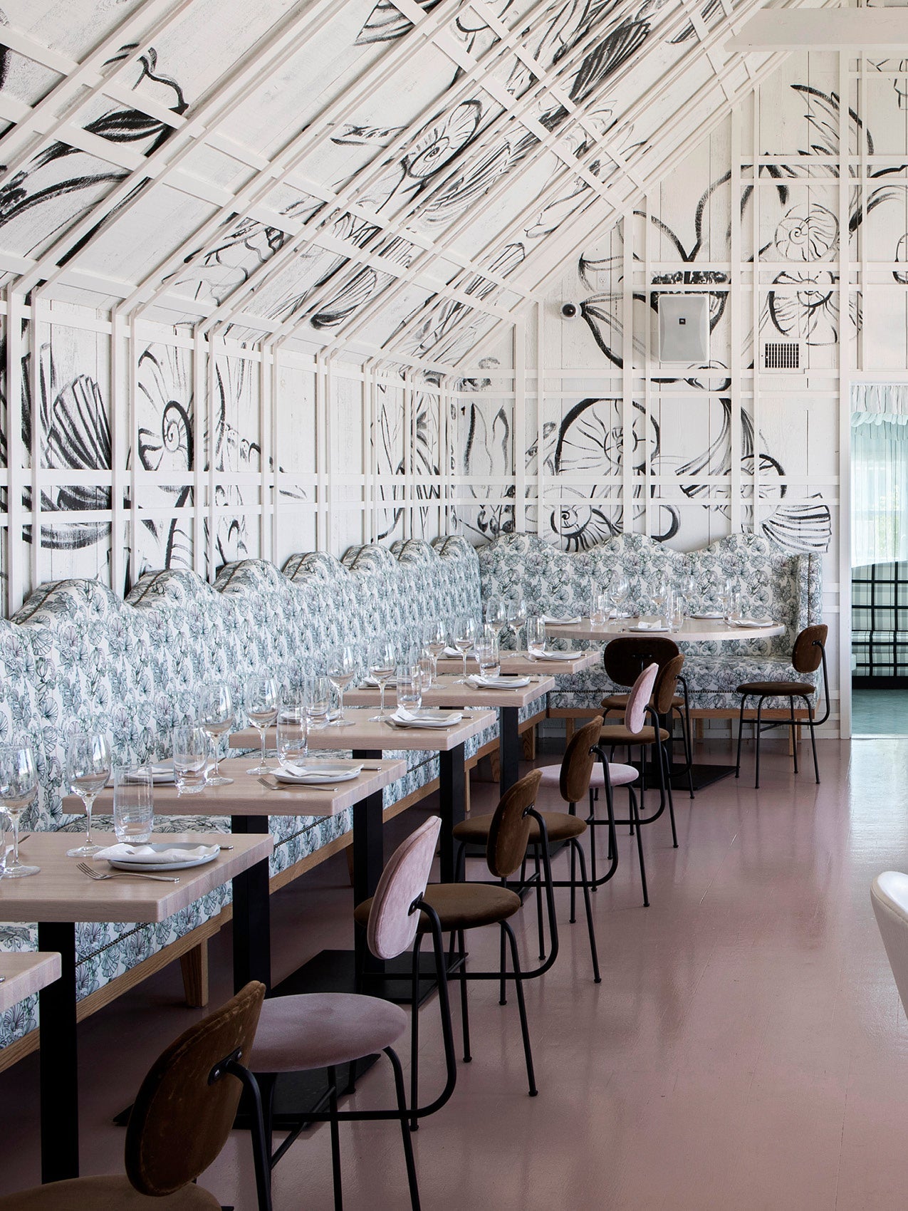 Bright dining room with floral-patterned banquette upholstery, pale wood tables, pink velvet chairs and white trellis wall