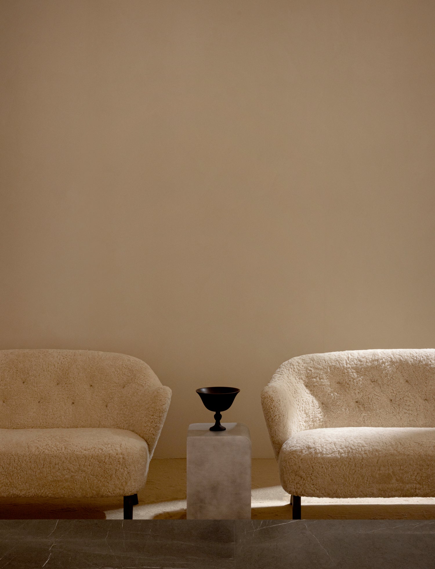 Pair of cream boucle armchairs flanking a small concrete pedestal topped with a black metal goblet-shaped bowl, neutral wall.