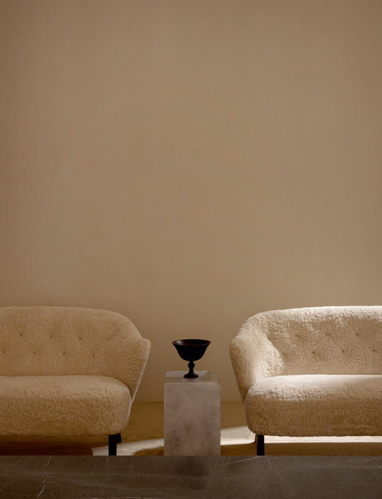 Pair of cream boucle armchairs flanking a small concrete pedestal topped with a black metal goblet-shaped bowl, neutral wall.