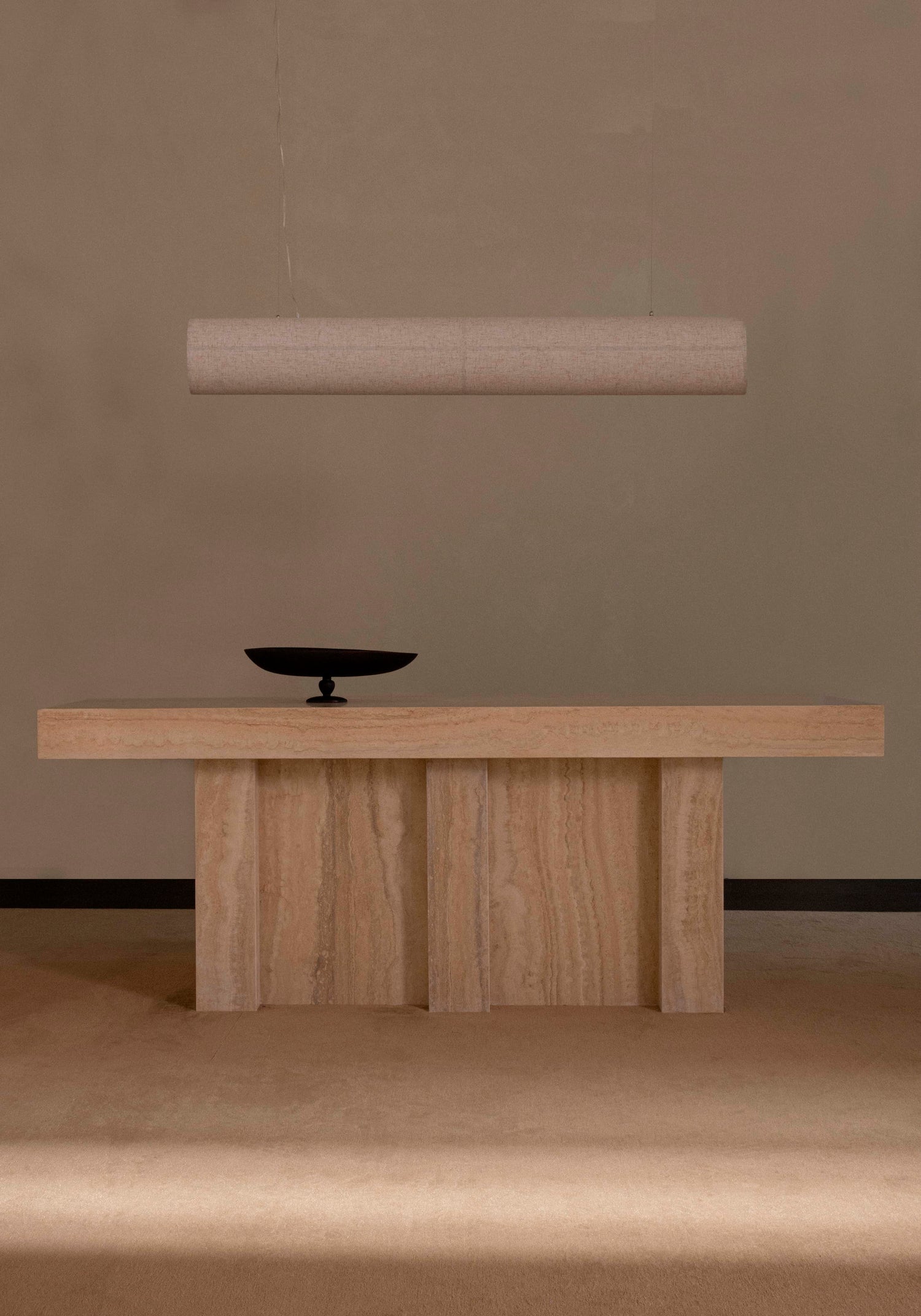 Minimal beige travertine console table with vertical panel legs, black shallow bowl on top and suspended cylindrical fabric
