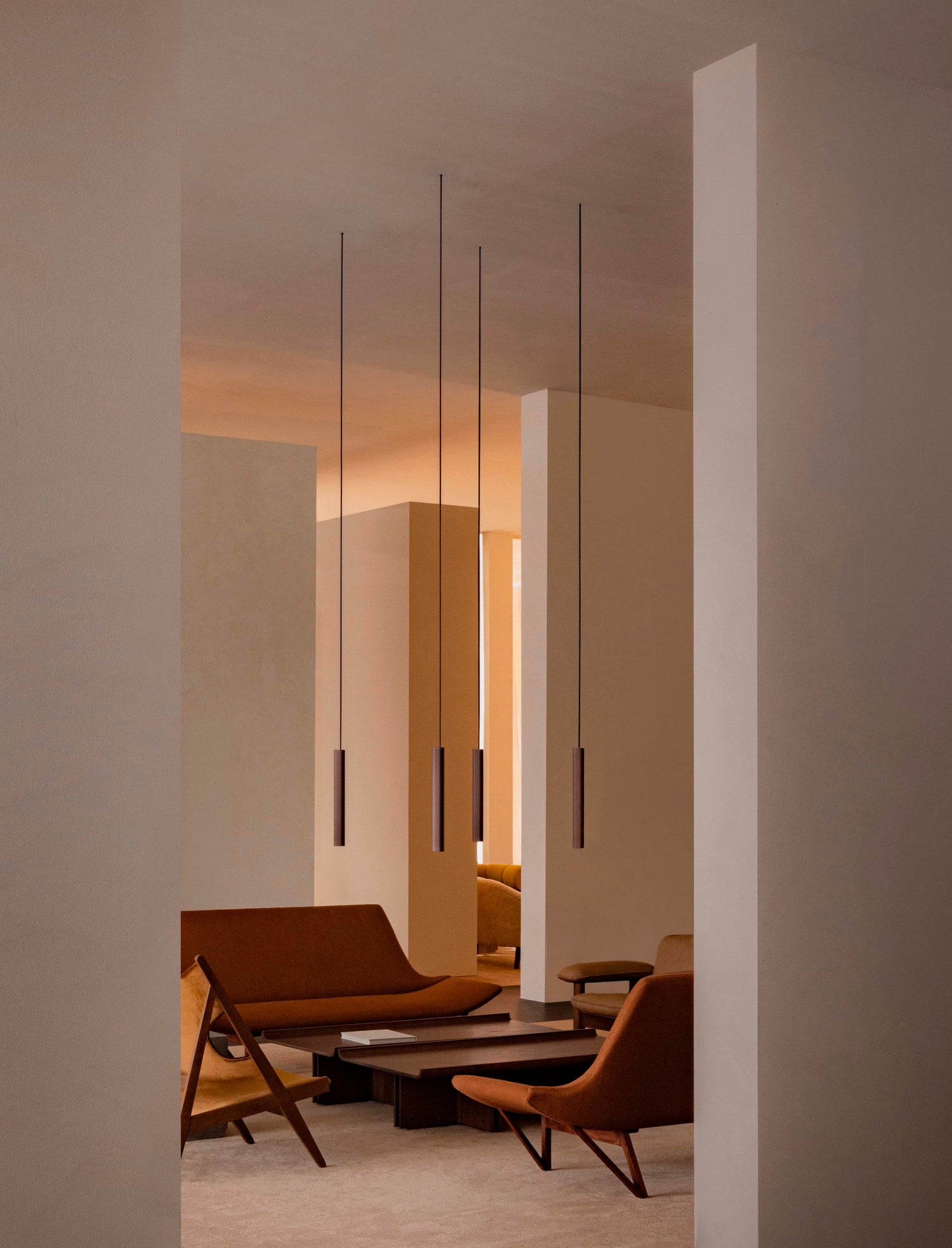 Minimalist interior with suspended slim brown pendant lights, warm beige walls and brown upholstered lounge chairs.