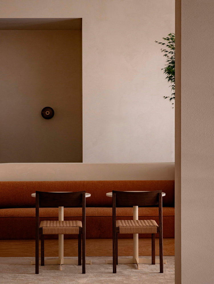 Minimal dining nook with two dark wood chairs, woven seats, round white tables and burnt orange banquette against beige