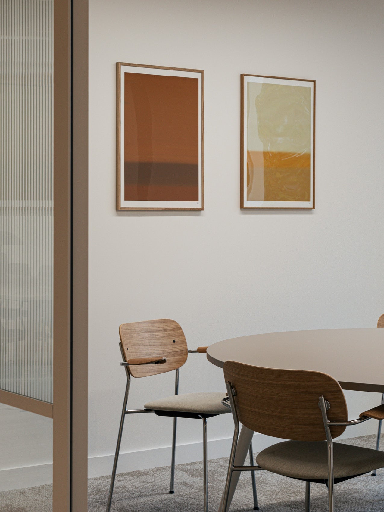 Minimal dining corner with round beige table and wooden chairs with metal legs, two framed abstract rust and ochre prints on