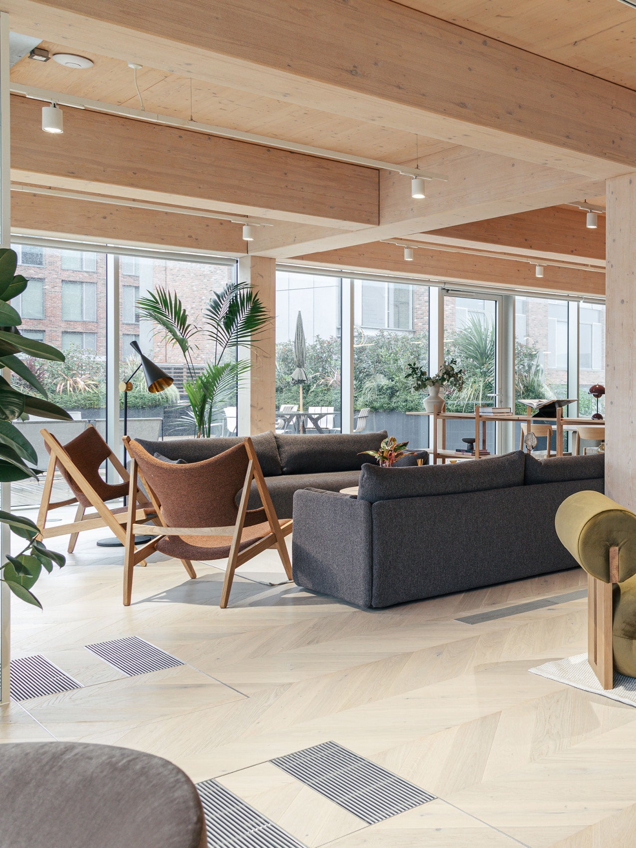 Open-plan lounge with grey fabric sofas, wooden-framed brown lounge chairs, light wood beams and large glass windows.