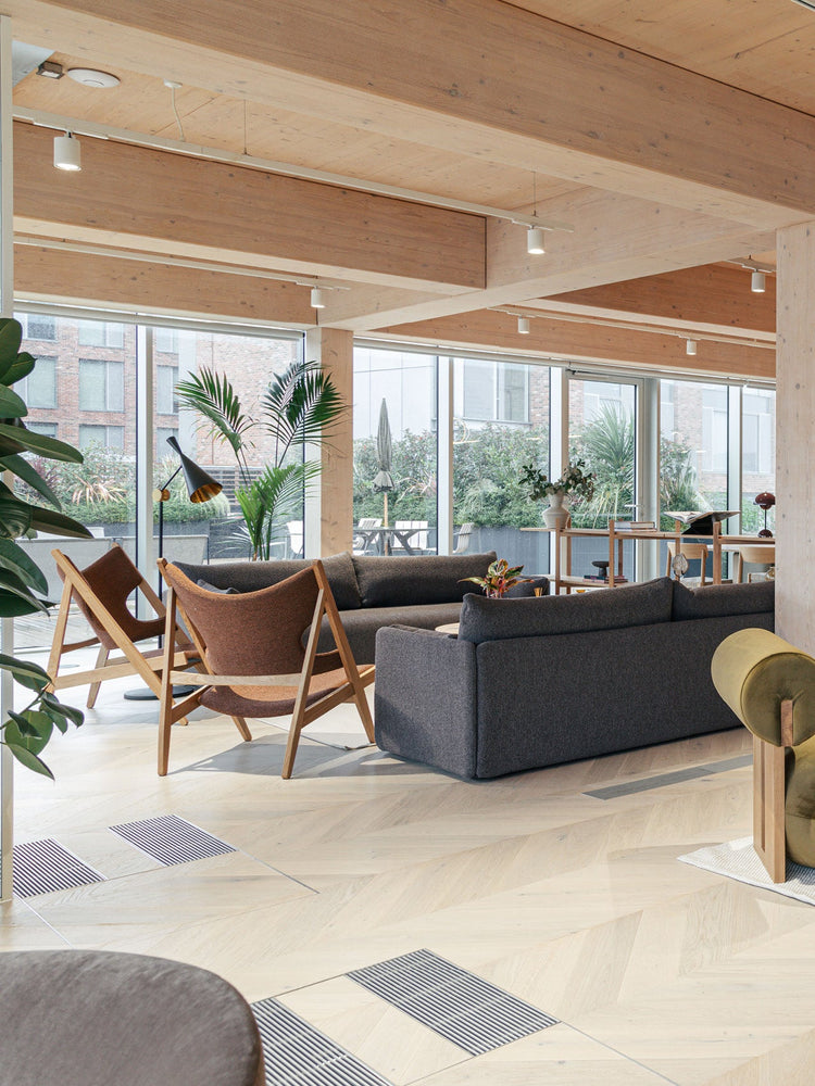 Open-plan lounge with grey fabric sofas, wooden-framed brown lounge chairs, light wood beams and large glass windows.