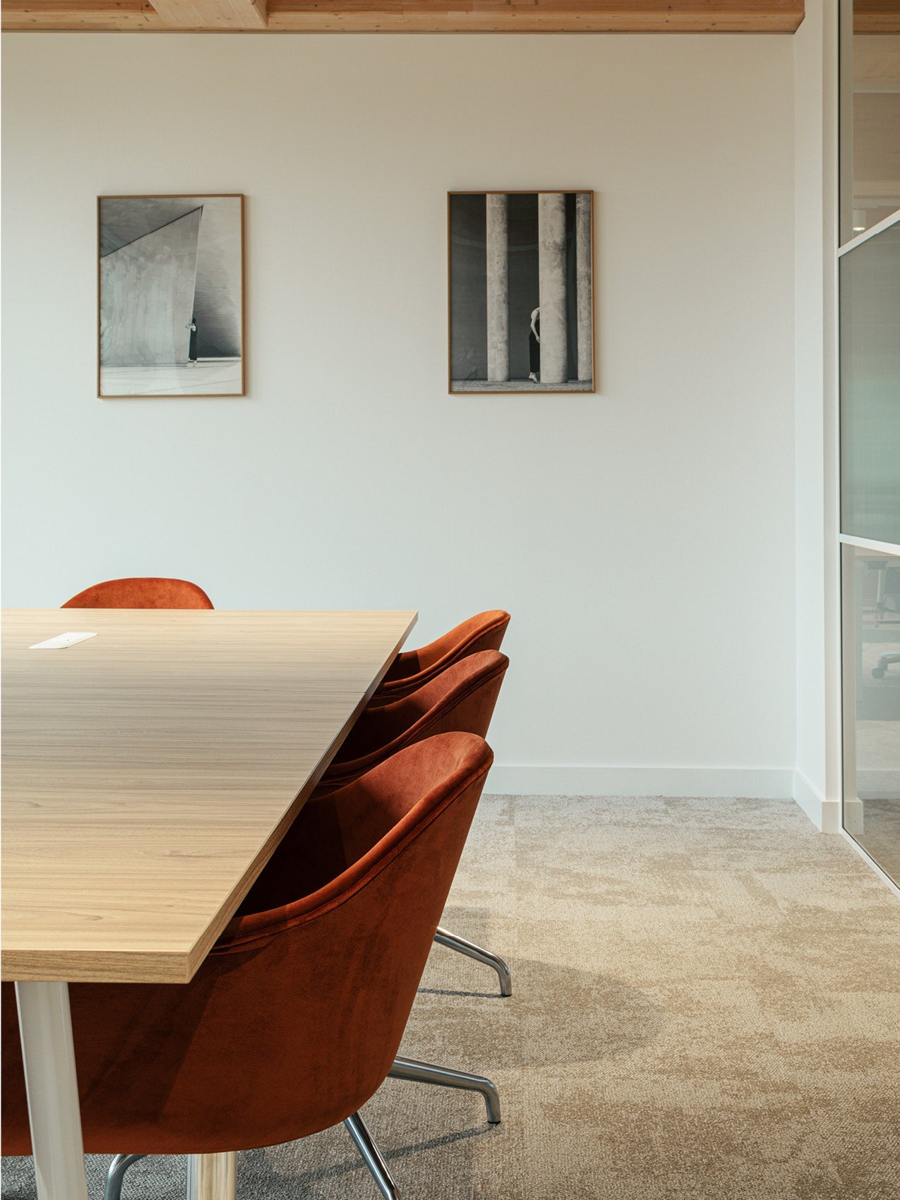 Minimal meeting room with light oak table and three rust-orange upholstered swivel chairs, neutral carpet and framed wall art