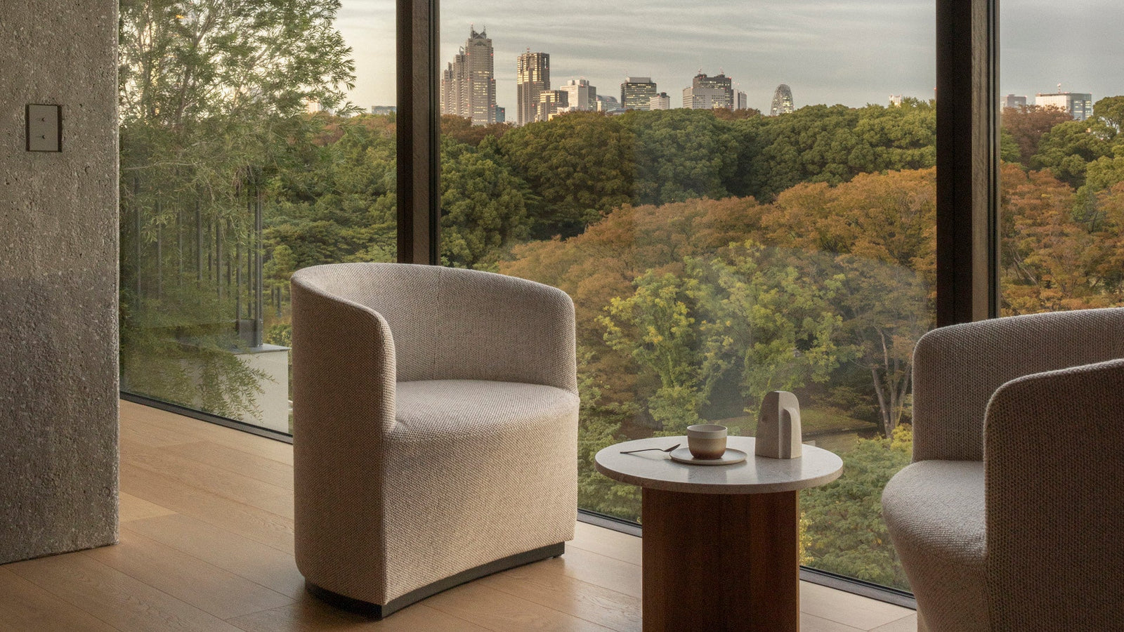 Beige boucle upholstered curved lounge chairs flanking a round wood side table with cup, large window showing green park and