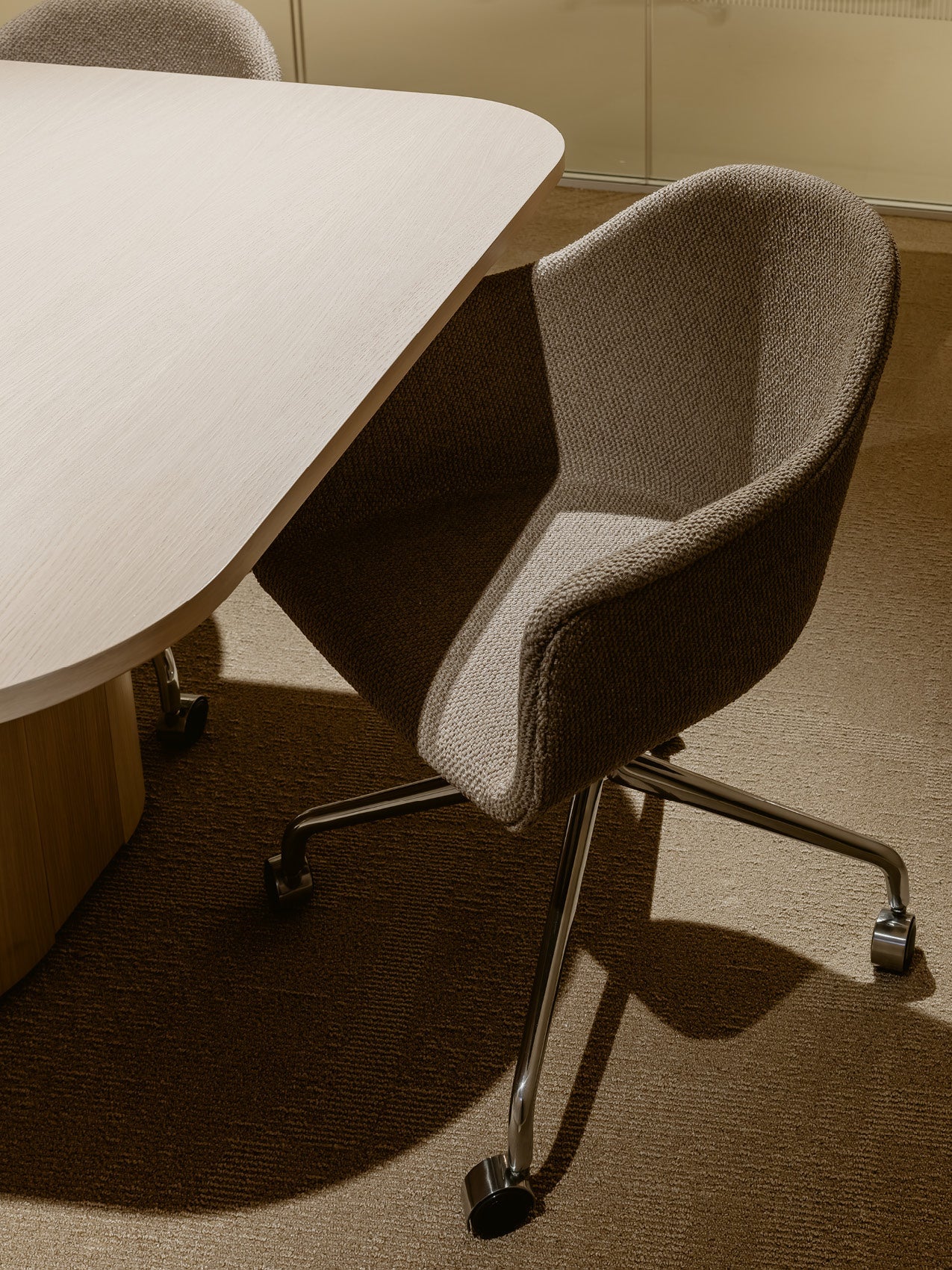 Beige upholstered swivel office chair with textured fabric and chrome base next to a light oak rounded-edge table.