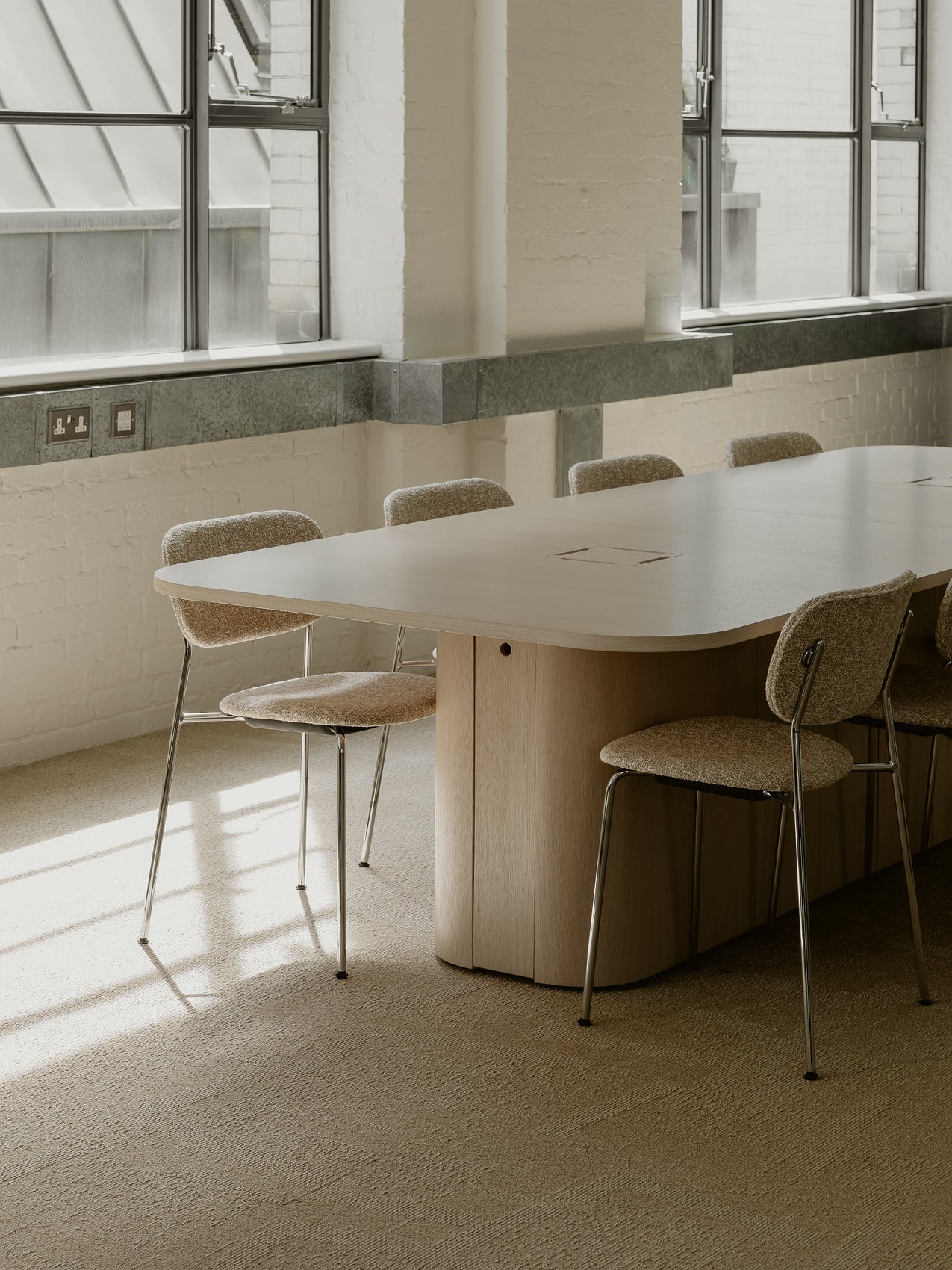 Light oak oval meeting table with rounded base and beige boucle chairs on chrome legs in sunlit room