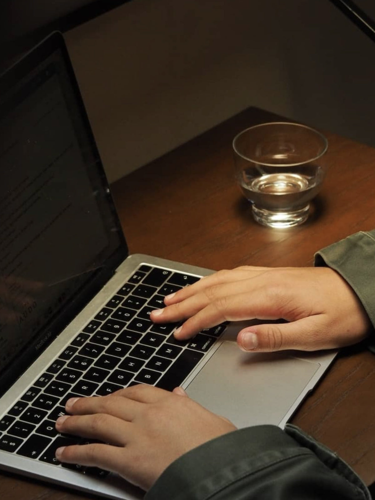 Hands typing on a laptop keyboard beside a glass of water on a wooden desk