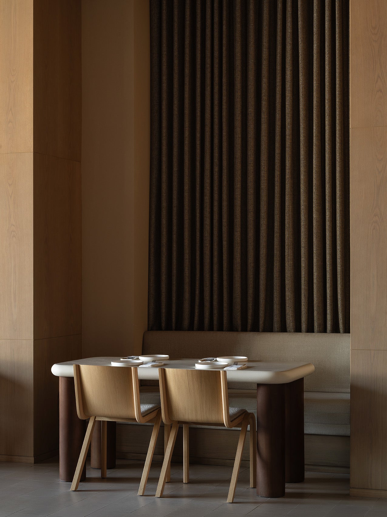 Minimal dining nook with light oak chairs, cream tabletop on dark cylindrical legs, and textured dark brown pleated wall