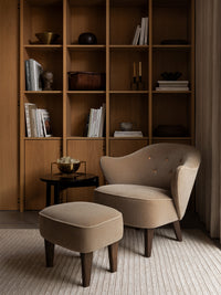 A beige Ingeborg Lounge Chair and Ingeborg Ottoman on a striped rug with a Kubus Bowl Centerpiece Large and wooden shelving.