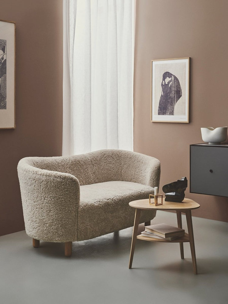 The Mingle Sofa in Sheepskin in a brown-walled room with a wooden side table, sheer curtains and framed modern wall art.