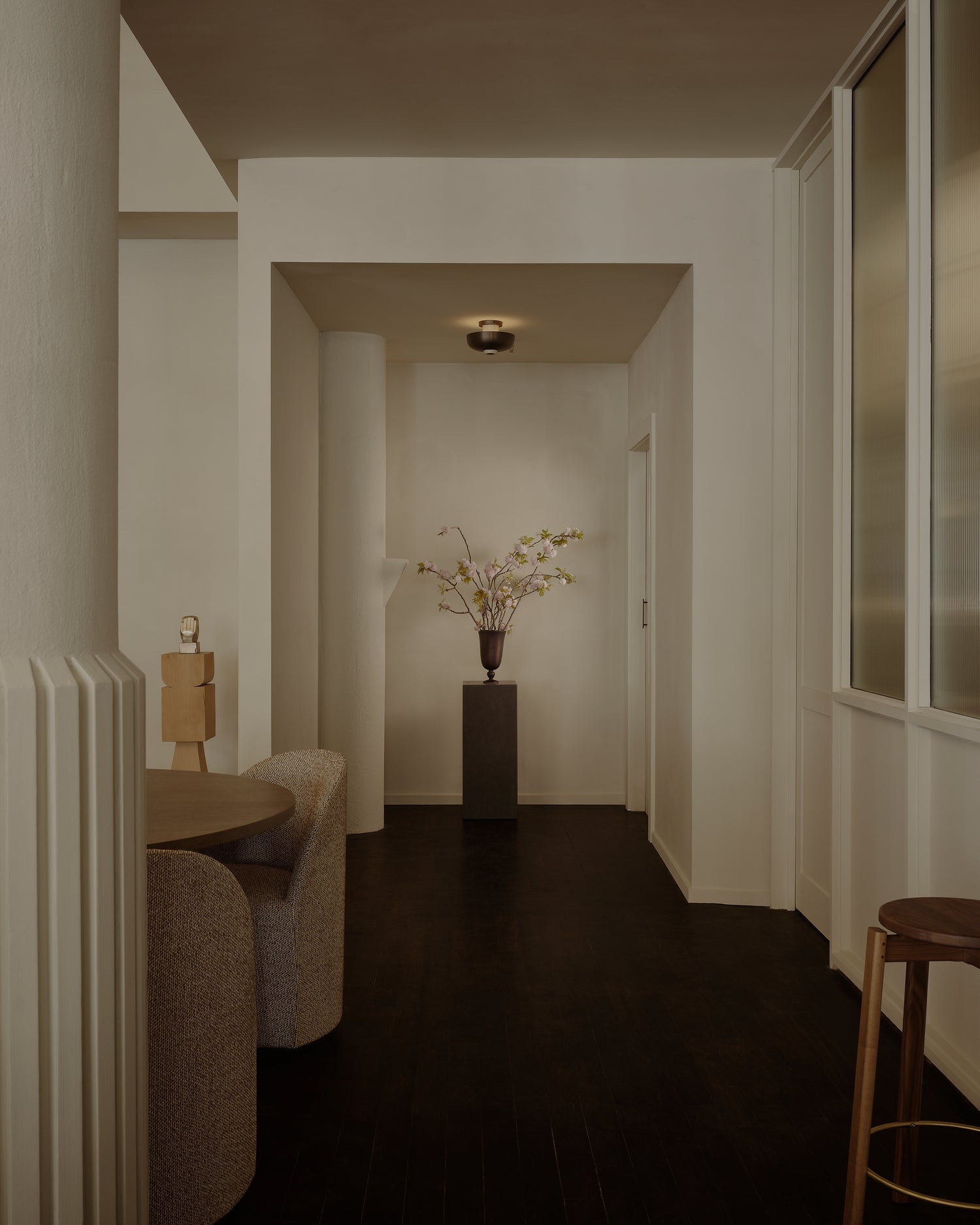 Minimal beige hallway with dark wood floor, central black pedestal holding a bronze vase of pale pink blossoms, ceiling light