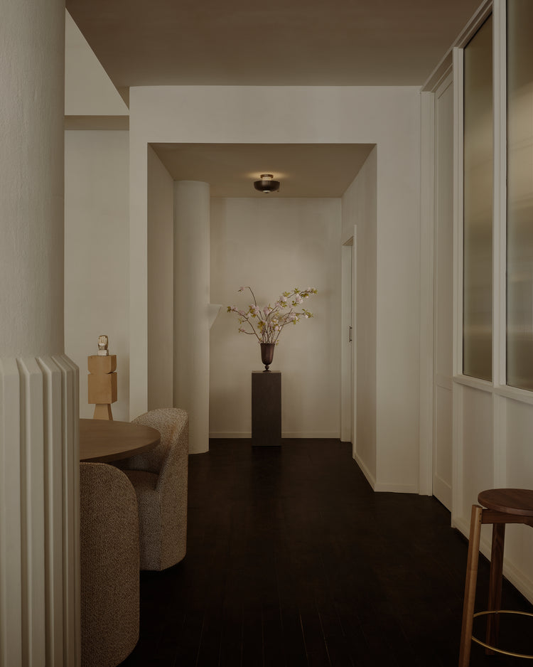 Minimal beige hallway with dark wood floor, central black pedestal holding a bronze vase of pale pink blossoms, ceiling light
