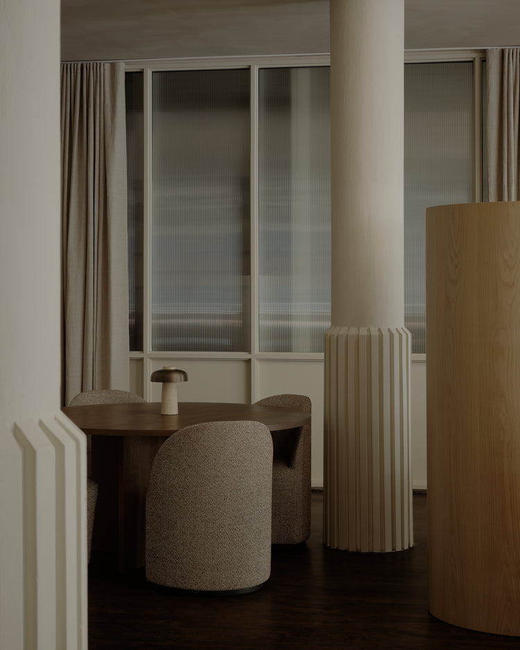 Minimal dining nook with round dark wood table, textured beige upholstered chairs, small cream ceramic lamp, fluted column