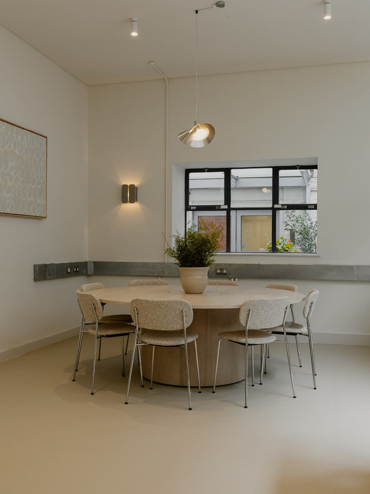 Round light wood dining table with six upholstered beige chairs, central ceramic planter, pendant and wall lights.