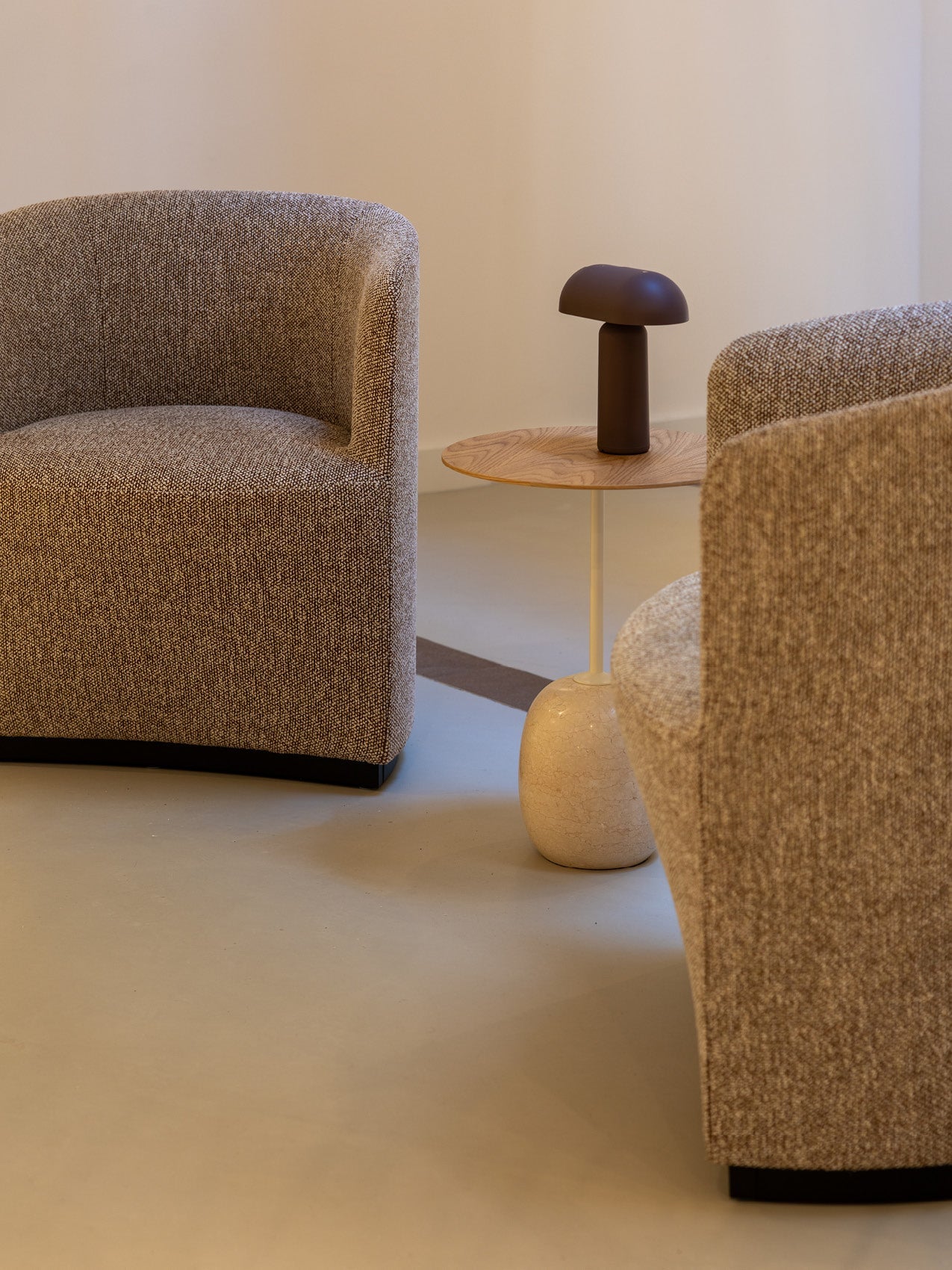 Pair of curved boucle armchairs in beige fabric with a small wooden side table topped by a matte brown mushroom table lamp