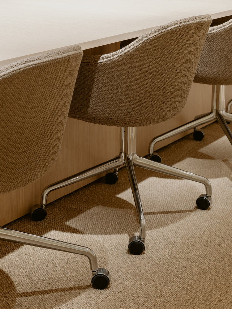 Three beige upholstered swivel chairs with chrome five-star bases and black castors tucked under a light wood table.