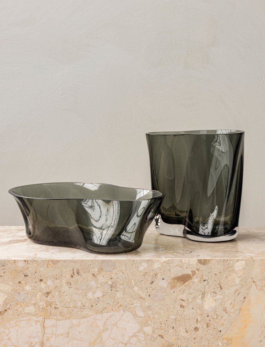 The Aer Vase and Aer Bowl in smoky grey glass, both with an organic, rippled shape, sit on a beige marble plinth.