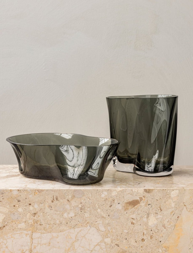The Aer Vase and Aer Bowl in smoky grey glass, both with an organic, rippled shape, sit on a beige marble plinth.