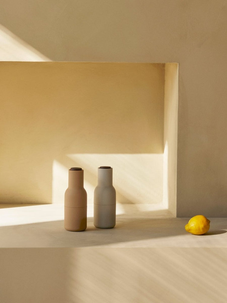 Two Audo Bottle Grinders, one beige and one light grey, on a stone surface in a sunlit, sandy-colored niche next to a lemon.