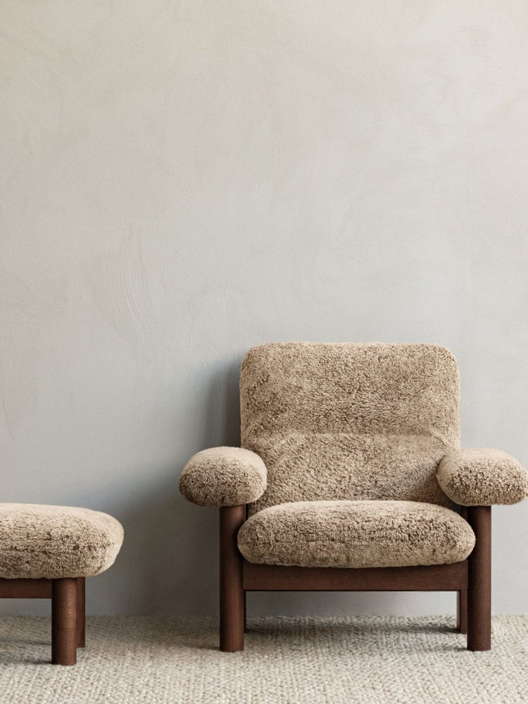 A Brasilia Lounge Chair and matching Brasilia Ottoman in brown sheepskin sit on a Gravel Rug against a light grey wall.