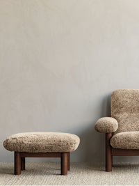 Close-up of the Brasilia Ottoman and Lounge Chair in brown sheepskin, placed on a light Gravel Rug against a grey wall.