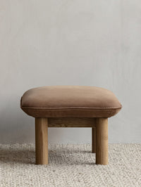 Front view of the Brasilia Ottoman, showcasing its brown textile cushion and robust natural oak legs on a gravel-colored rug.