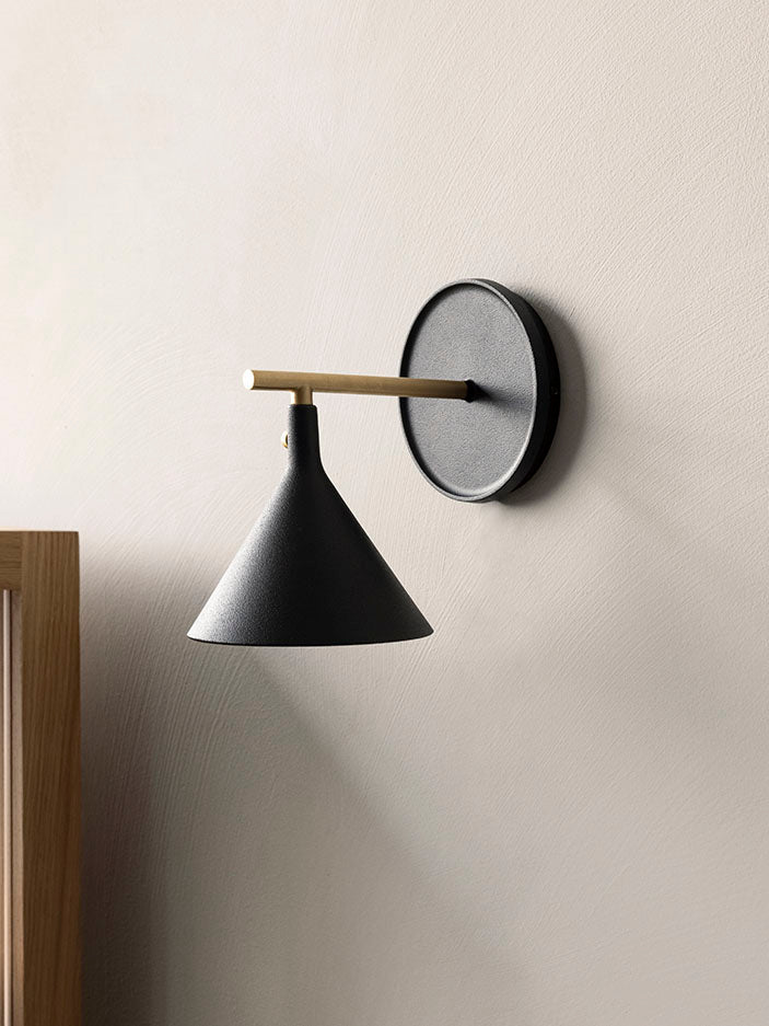 The black Cast Sconce Wall Lamp is shown mounted on a textured off-white wall, with a brass arm and a cone-shaped shade.