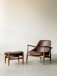 Elizabeth Lounge Chair and Elizabeth Ottoman in brown leather with walnut frames, set against a plain wall on a gray floor.